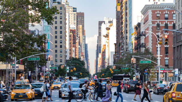 Busy crowds of people walk across 3rd Avenue in front of rush hour traffic in the East Village neighborhood of Manhattan in New York City.