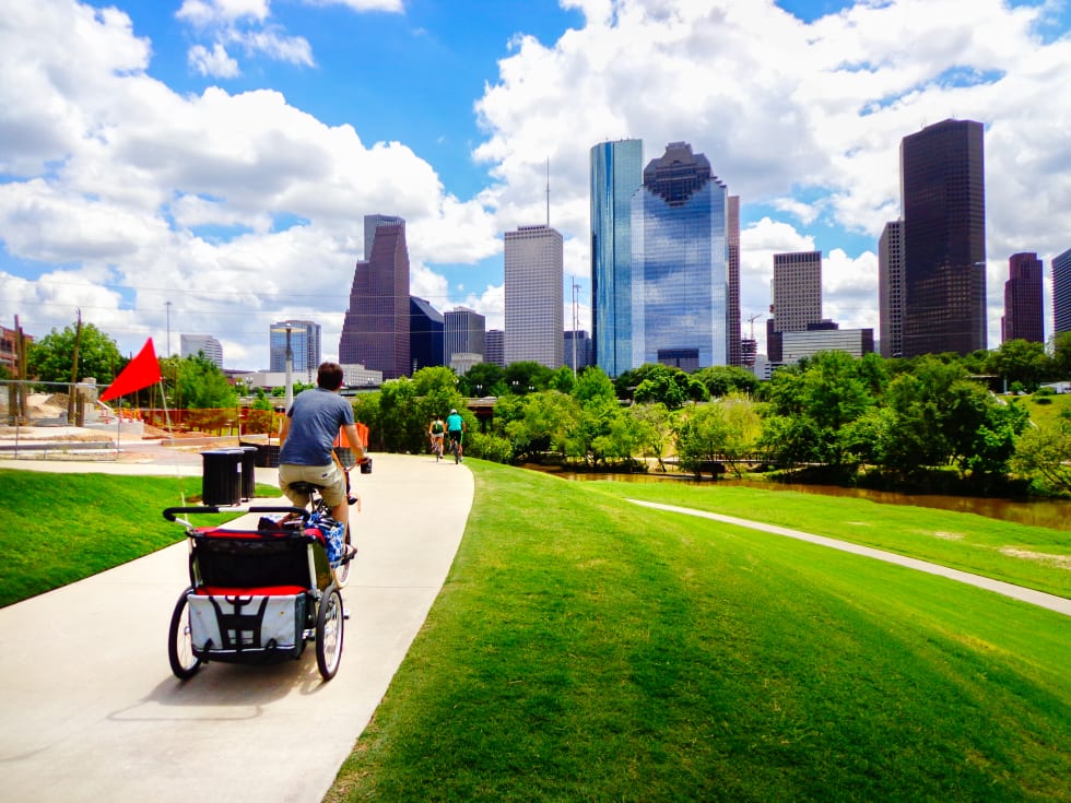  Young man riding bike with baby carriage attached on paved trail in Houston park (view of river and skyline of downtown Houston) 