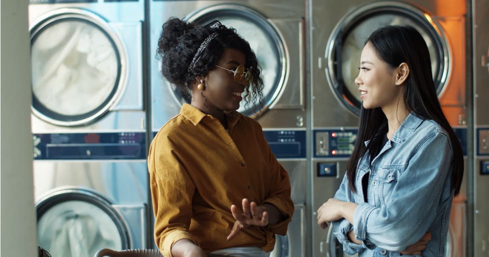 Stylish young girls best friends talking and sharing secrets while standing in public laundromat. 
