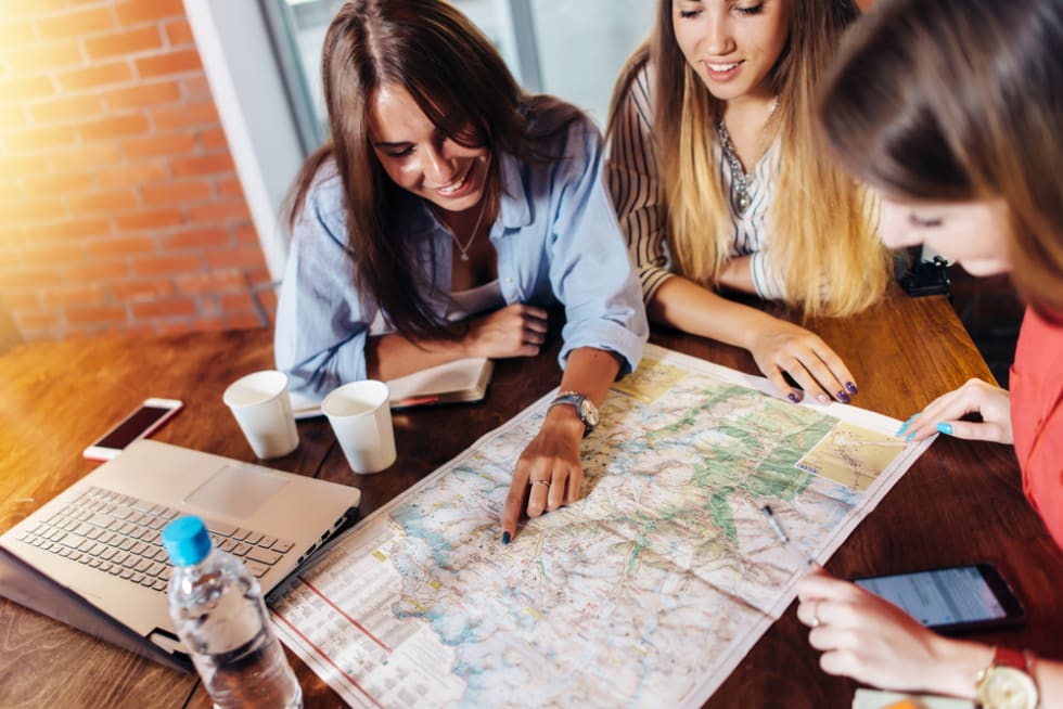 Smiling female friends sitting at desk planning their vacation looking for destinations on map