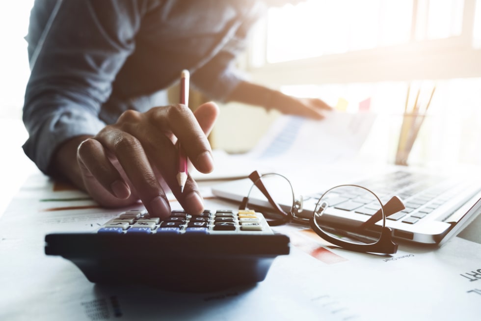 Close up of businessman or accountant hand holding pen working on calculator to calculate business data, accountancy document and laptop computer at office, business concept