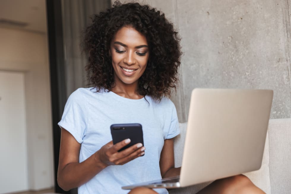 Woman using mobile phone while sitting with laptop computer on a couch at home