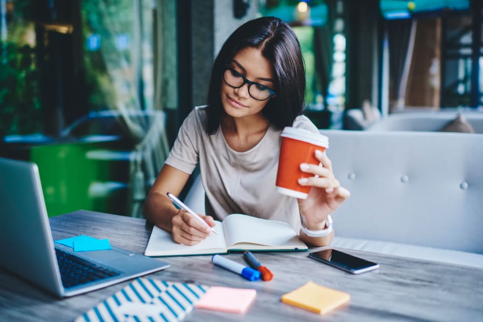 Concentrated female student writing in notebook while learning with cardboard coffee cup in cafe