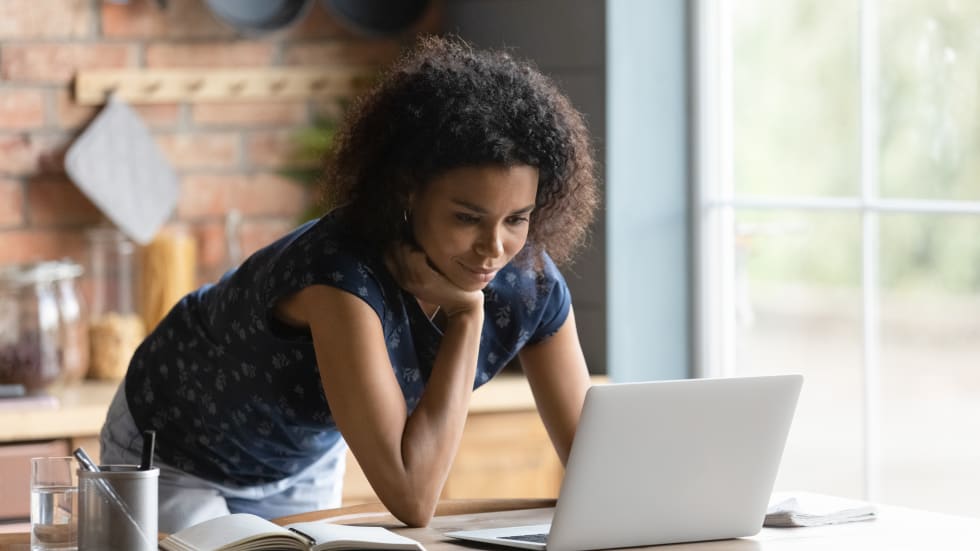 woman using laptop, standing at table in kitchen, working on research project or studying online at home, watching webinar on computer screen, reading news - free furniture pick-up