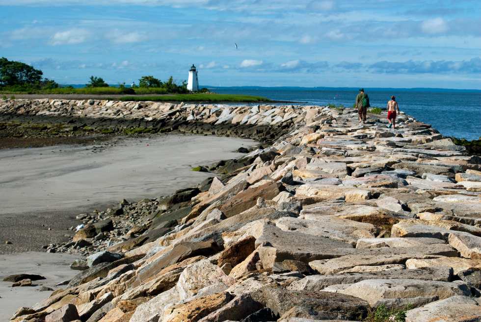 Young couple hiking along rocky breakwater towards Black Rock Harbor light connecting to Fayerweather Island in Bridgeport, Connecticut. 