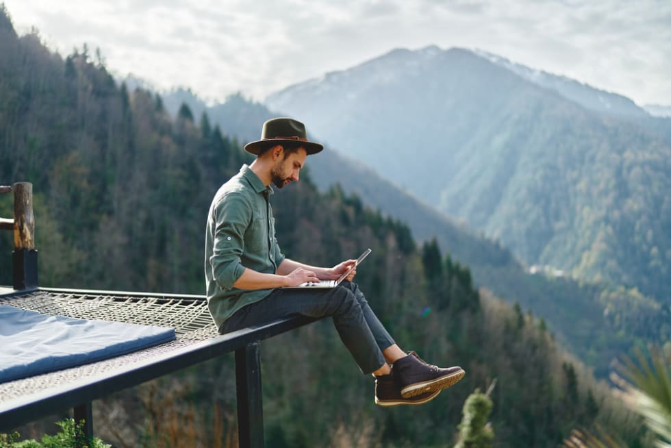 Young man freelancer traveler wearing hat anywhere working online using laptop and enjoying mountains view