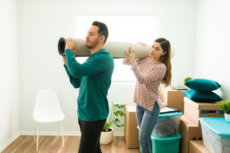 Side view of a young woman and man holding a rolled carpet together. Boyfriend and girlfriend carrying a rug to put on their new bedroom