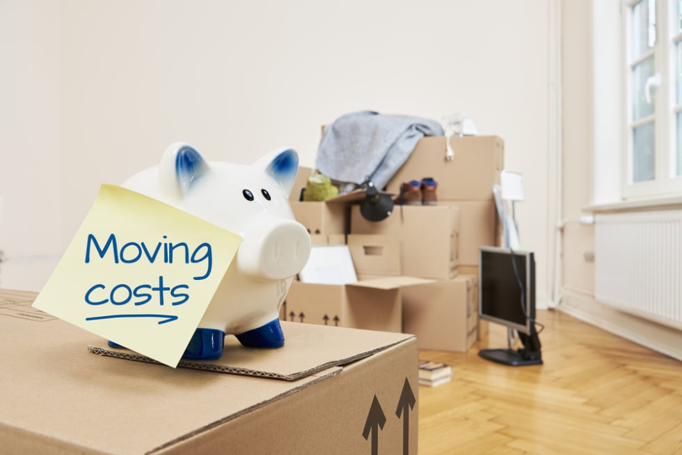 On a moving box there is a piggy bank on which a yellow piece of paper sticks. On the note is moving coats. In the background is a stack of packed moving boxes