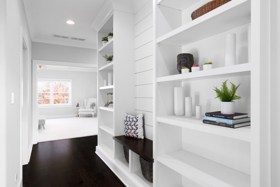 A white, built-in shelving in the hallway with shiplap, decorations on the shelves, a wooden bench, and looking towards a bright bedroom.