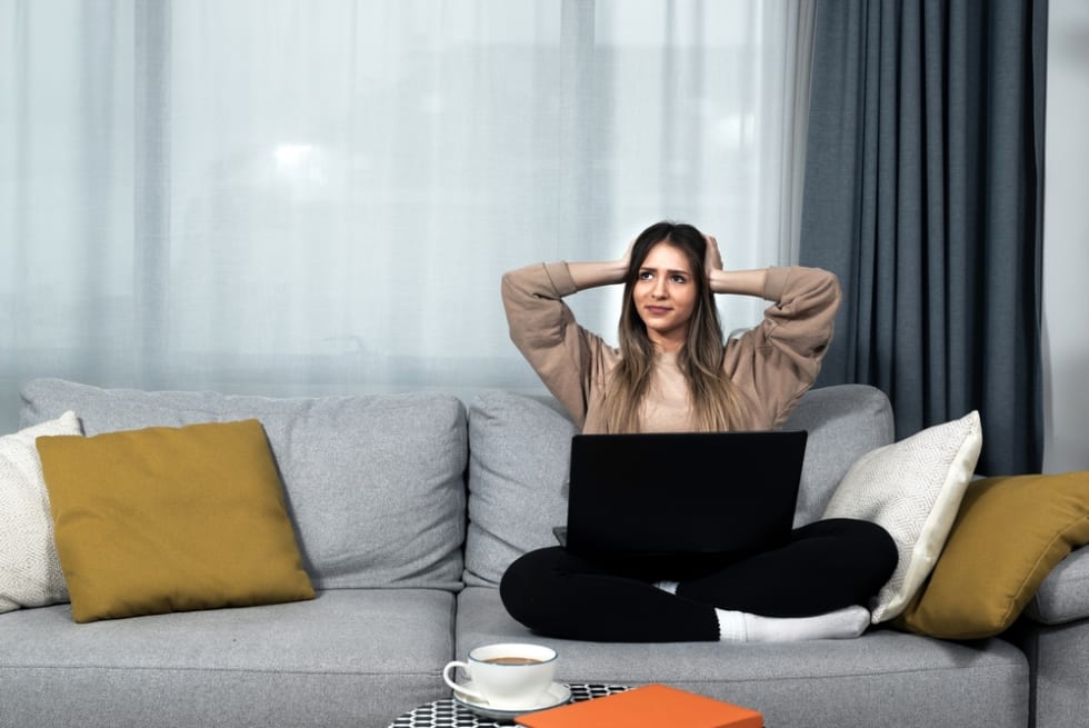 Young student or business woman sitting at home covering her ears with hands because she cant study or work on her laptop computer from loud noise coming from apartment above.