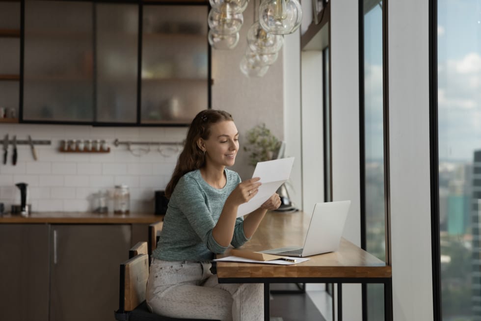 Happy young woman reading paper letter at home workplace with laptop, getting good news, bank or insurance notice. 