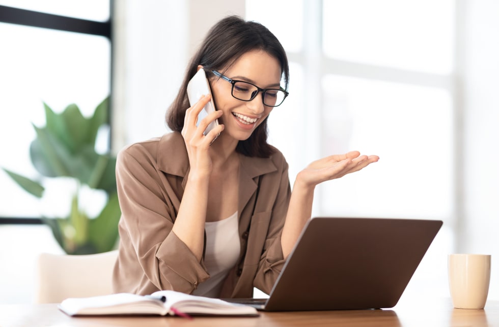 Happy young woman in glasses calling her friend, talking and asking about something, using laptop