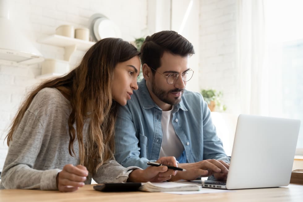 Focused young couple calculating bills, discussing planning budget together, serious wife and husband looking at laptop screen, using online banking services and calculator, checking finances - rent an apartment remotely