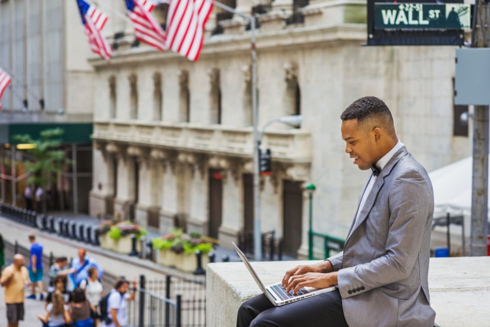  college student siting on Wall Street, reading, working on laptop computer.